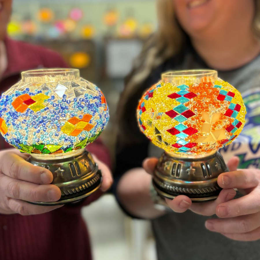 Two people holding handmade mosaic glass globes with ornate bronze bases in a brightly lit indoor setting.